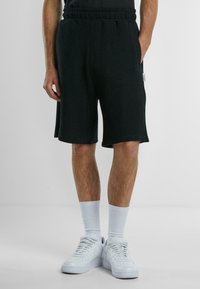 Svarta ribbade shorts med elastisk midja, sidfickor och en avslappnad passform, i kombination med vita sneakers och ljusblå strumpor.