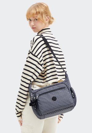 Une jeune personne avec les cheveux blonds courts porte un pull rayé et tient un sac à bandoulière noir et blanc à motifs avec un porte-clés en peluche.