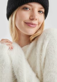 Witte, fluffy trui met een zachte textuur, in combinatie met een zwarte ribgebreide beanie. Het model heeft blond haar en houdt de trui voorzichtig vast.