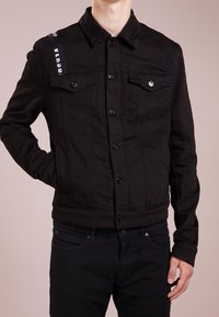 Giacca in denim nero con collo a camicia, tasche frontali e chiusura con bottoni. Presenta un dettaglio a righe sulla spalla. Design aderente.