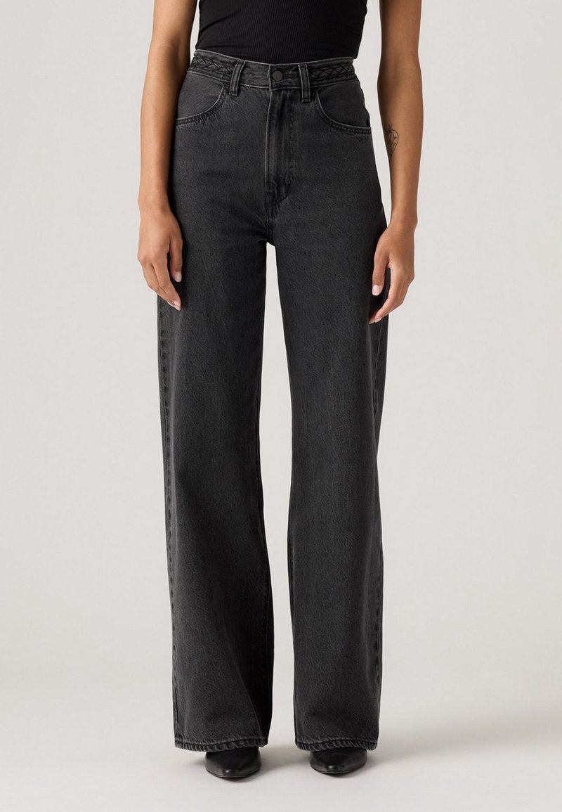 Femme portant un jean noir taille haute à jambes larges et des chaussures noires, debout devant un fond clair uni.