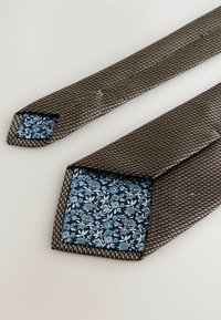 Smeđa tkana kravata s cvjetnim plavim i bijelim detaljima na stražnjoj strani. Kravata ima teksturiranu površinu i dizajn s oštrim vrhom.