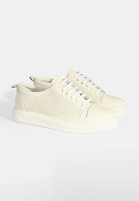 Beige Sneaker mit einer glatten Lederkappe und perforierten Seitenteilen, ausgestattet mit weißen Schnürsenkeln, einer flachen Gummisohle und einer Zugschlaufe an der Rückseite.