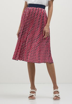 Donna che indossa una gonna midi plissettata con un motivo geometrico rosa, rosso e nero, una maglietta bianca e sandali con tacco alto bianchi e a strisce.