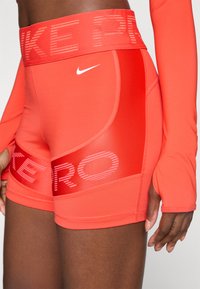 Zatvoren kadar osobe koja nosi svijetlu crvenu Nike Pro kratku hlače i odgovarajući dugih rukava majicu s otvorima za palčeve, prikazujući Nike logo i teksturirani pojas.