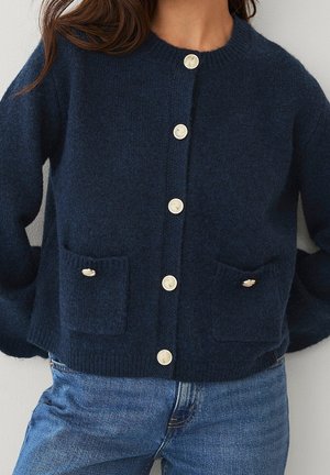 Femme portant un cardigan bleu marine boutonné avec des poches plaquées, assorti à un jean bleu, montrée des épaules aux hanches.