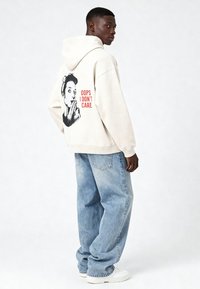 Béžová mikina s kapucňou a grafickou potlačou na zadnej strane zobrazujúcou postavu a text "OOPS I DON'T CARE." Voľné modré rifle z denimu a biele tenisky.