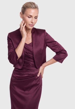Woman with blonde hair in a sleek updo wears a plum satin dress and matching cropped jacket, posing thoughtfully with one hand on her face.