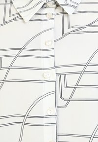Blusa bianca con motivo lineare nero, caratterizzata da un colletto a punta, chiusura frontale con bottoni rotondi e tessuto liscio e leggero.