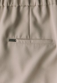 Beige stof met een gladde textuur. Bevat een rechthoekige zak met een metalen accentdetail, met zichtbare stiksels langs de randen.