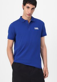 Blauwe polo gemaakt van katoen, met een klassieke kraag, korte mouwen en een wit logo op de linkerborst. Zachte textuur, relaxte pasvorm.