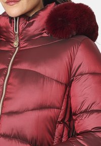 Chaqueta acolchada roja con un acabado brillante y acolchado, que presenta un borde de pelaje en la capucha y un detalle de cremallera dorada. Textura suave y diseño acolchado.