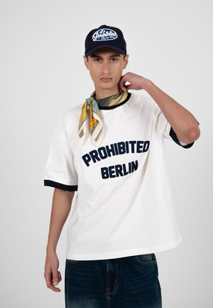 Giovane uomo che indossa una maglietta bianca con la scritta "PROHIBITED BERLIN", un cappellino blu scuro con scritta, una sciarpa gialla con motivi e jeans scuri, posando davanti a uno sfondo neutro.