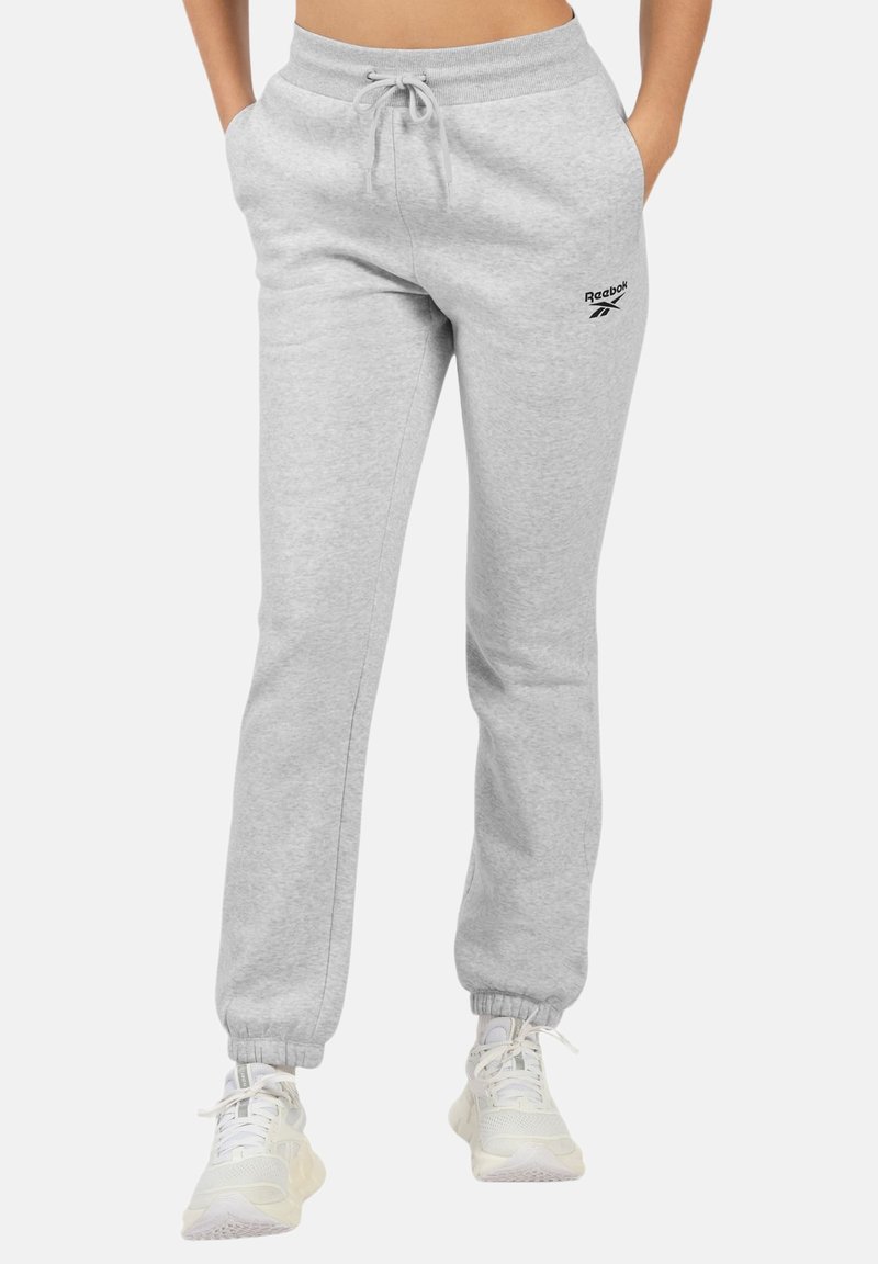 Pantalon de survêtement Reebok gris clair avec taille à cordon de serrage et poignets élastiques, porté avec des baskets blanches de sport, mains dans les poches.