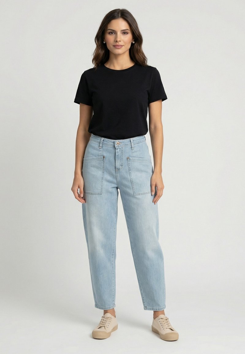 Donna in piedi che indossa una maglietta nera a maniche corte, jeans a vita alta azzurri chiaro con tasche frontali e scarpe da ginnastica beige.