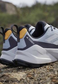 Ζευγάρι γκρι και λευκά παπούτσια trail running Terrex με μαύρες σόλες και κίτρινες λεπτομέρειες στη φτέρνα σε βραχώδες έδαφος έξω.