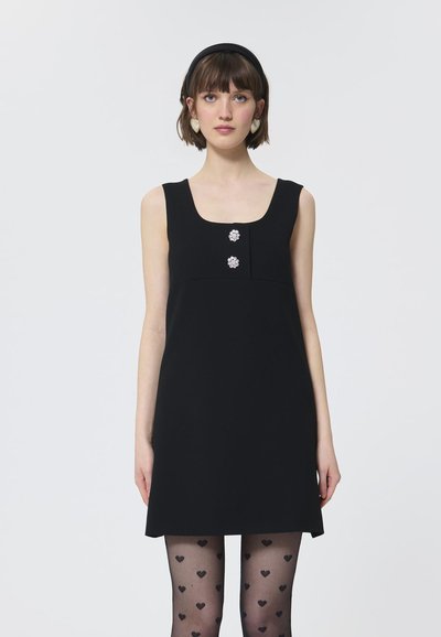 Robe noire sans manches avec un décolleté carré, ornée de deux embellissements en forme de fleurs argentées. Le mannequin porte des collants noirs à motifs de cœurs.