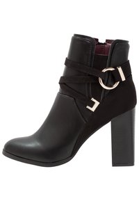 Bota negra hasta el tobillo con parte superior de cuero liso, acentos de tiras decorativas, punta redonda y un tacón grueso y alto. Presenta un detalle de hebilla dorada.