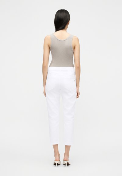 Γυναίκα με μακριά σκοτεινά μαλλιά φοράει μπεζ αμάνικο τοπ, λευκό cropped τζιν και λευκά σανδάλια με τακούνι, στέκεται μπροστά σε απλό φωτεινό φόντο.