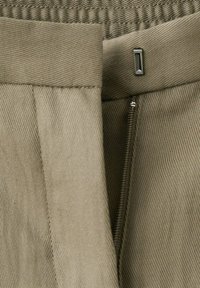 Fermeture à agrafes avec fermeture éclair partiellement ouverte sur un tissu en maille côtelée beige, montrant les coutures de la ceinture et les détails de texture.