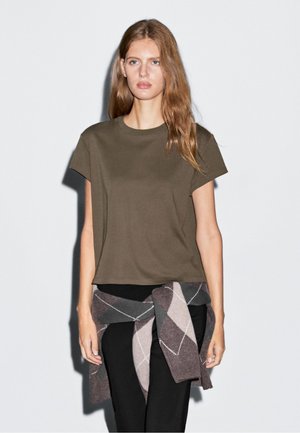 Olive green cotton t-shirt with a relaxed fit and short sleeves, paired with black pants and a brown argyle knit sweater tied around the waist.