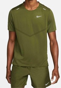 Hombre vestido con una camiseta deportiva Nike de color verde oliva y pantalones cortos a juego, con logotipos blancos de Nike, de pie frente a un fondo blanco.