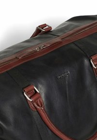 Black leather duffel bag with brown leather accents, a top zipper, and adjustable straps. Features embossed logo and smooth texture.