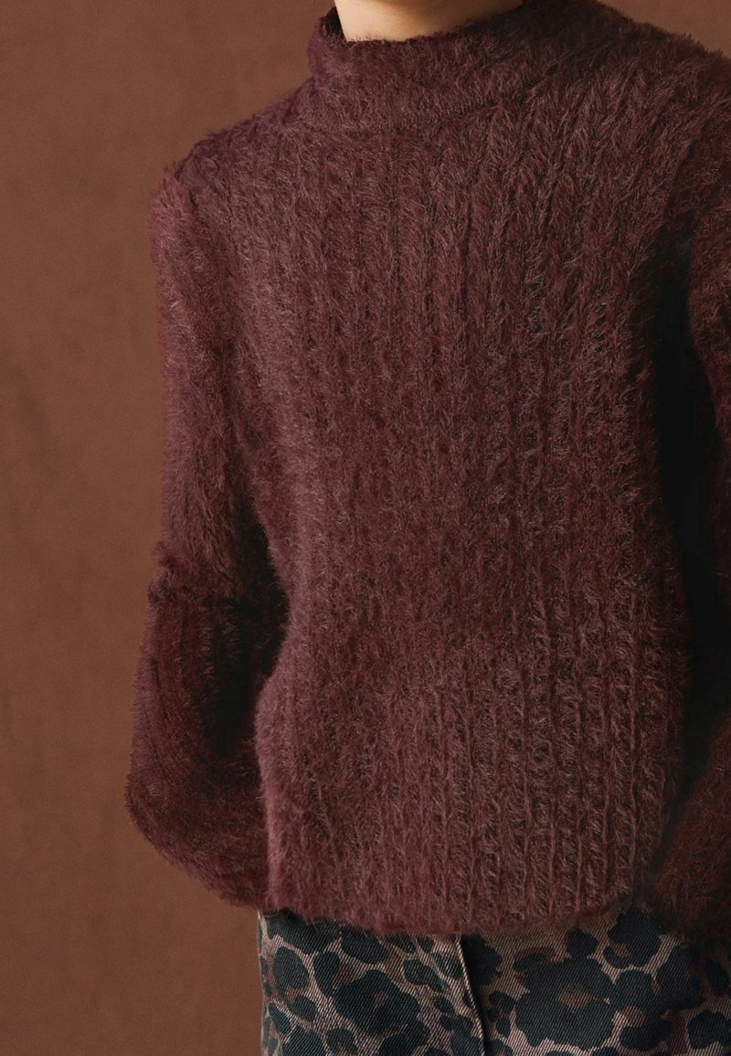 Gros plan d'une personne portant un pull torsadé en laine pelucheuse de couleur bordeaux foncé et un pantalon à motif léopard sur un fond marron.