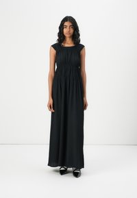 Svart maxi-klänning med plisserad textur, korta ärmar och figurnära midja. Modellen bär svarta pumps med spetsig tå. En enkl vit bakgrund.