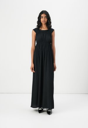 Rochie maxi neagră cu textură plisată, mâneci scurte și talie ajustată. Modelul poartă pantofi negri cu vârf ascuțit. Fundal alb simplu.