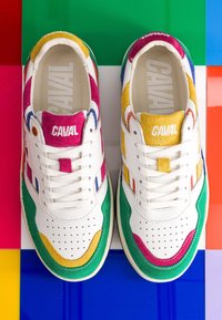 Paire de baskets blanches avec des touches de daim vert, jaune, rose, bleu et le logo « CAVAL » sur la languette, posée sur un fond de carreaux multicolores.