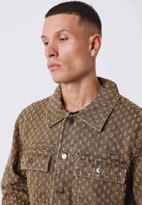 Homme aux cheveux courts portant une veste marron texturée avec des boutons en métal et une boucle d'oreille en diamant, regardant vers le bas et sur le côté.