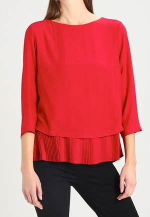 Blouse rouge avec un col rond et des manches trois-quarts. Présente une couche plissée au niveau de l'ourlet. Texture de tissu lisse, design décontracté.