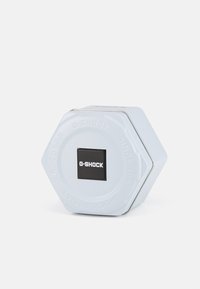 Boîte en métal hexagonale G-SHOCK en bleu clair avec une étiquette carrée noire. Texture lisse, fermoir en métal et détail de la marque en relief sur le couvercle.