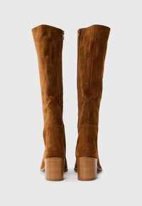Bottes en suede marron au-dessus du genou avec une forme contoured, un détail de fermeture éclair sur le côté et un talon en bois empilé. Texture lisse avec une finition mate.