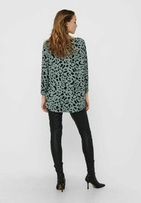 Tunique vert clair avec motif abstrait noir, manches trois-quarts, coupe décontractée, associée à un pantalon en cuir noir ajusté et des bottes à talons.