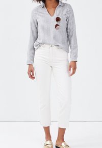 Chemise rayée à col en V, associée à un jean blanc court. La tenue comprend des chaussures dorées à enfiler et des lunettes de soleil.