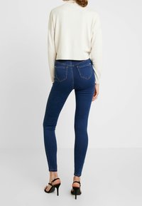 Jeans ajustados en denim azul oscuro con cintura alta, que cuentan con bolsillos traseros y una textura suave, combinados con un suéter corto crema.