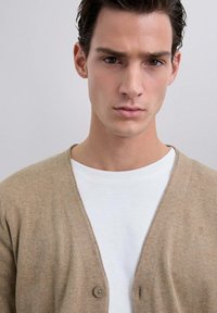 Cárdigan beige con escote en V y botones frontales, superpuesto sobre una camisa blanca. Textura suave, tela blanda, diseño minimalista, look casual.