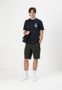 T-shirt en coton bleu marine avec logo blanc, associé à un short cargo vert olive. Tenue complétée par des baskets blanches et un petit sac noir.