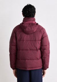 Bordeauxfarbene Daunenjacke mit Kapuze, die über gesteppten Bereiche und eine glatte Textur verfügt. Der Rücken zeigt elastische Bündchen und einen abgerundeten Saum.
