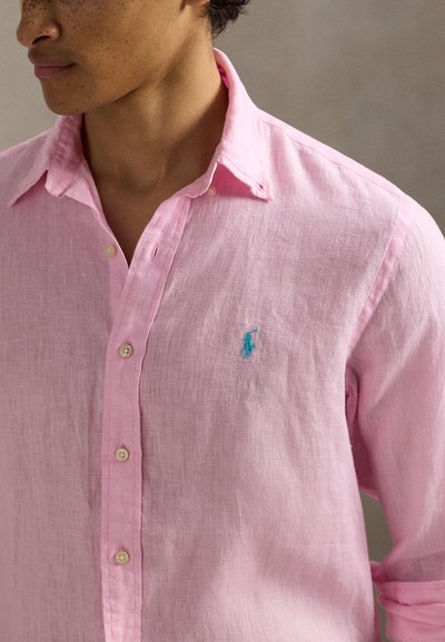 Férfi világos rózsaszín vászongombos ingben, bal mellkason kék hímzett logóval, nyitott gallérral, felhajtott ujjakkal.