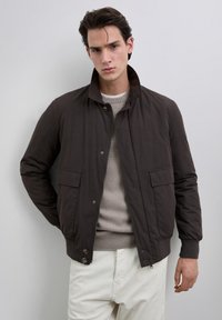 Joven con una chaqueta marrón oscura sobre un suéter beige y pantalones blancos, de pie contra un fondo gris claro liso.