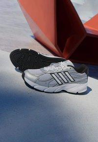 Biele športové topánky s sieťovinou, šedými a čiernymi akcentmi, textúrovanou čiernou gumovou podrážkou a tromi paralelnými čiernymi pruhmi po boku.