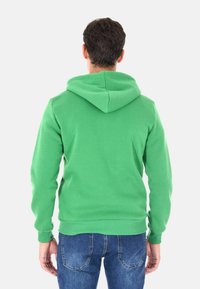 Felpa verde con cappuccio regolabile, bordi a costine e una texture liscia, abbinata a jeans blu e uno sfondo semplice.