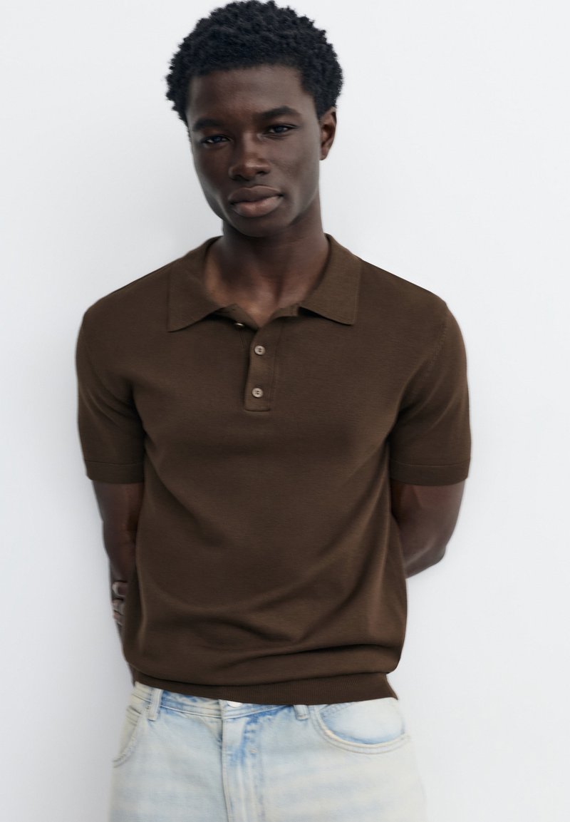 Jeune homme aux cheveux courts et bouclés, portant un polo marron à manches courtes et un jean bleu clair, debout devant un fond blanc.