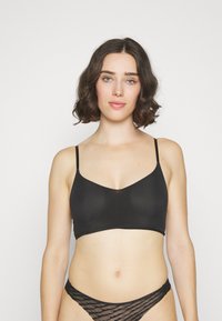 Crna bralette s podesivim trakama, glatki materijal, V-izrez i minimalni šavovi, u kombinaciji s prozirnim crnim teksturiranim donjim rubljem.