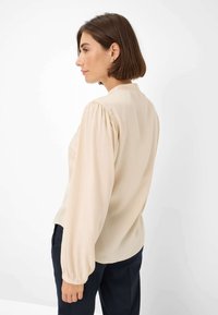 Beige blouse with puffed long sleeves, smooth fabric, rounded hem, and a high neckline. Worn with dark trousers, showing a side view.
