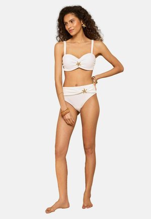 Vrouw met krullend haar die een witte, getextureerde bikini draagt met gouden zeesterren als versiering, poserend op blote voeten tegen een effen lichte achtergrond.