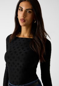 Czarna bluzka z długim rękawem o fakturowanym wzorze, z kwadratowym dekoltem i dopasowanym krojem. Noszona z wysokim stanem dżinsów.